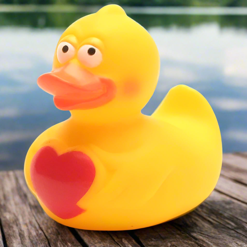 A yellow rubber duck with a red heart-shaped detail on its back, positioned on a wooden surface with a blurred background.
