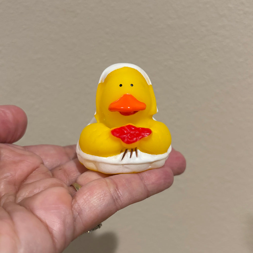 Hand holding a small yellow rubber duck toy against a beige wall.
