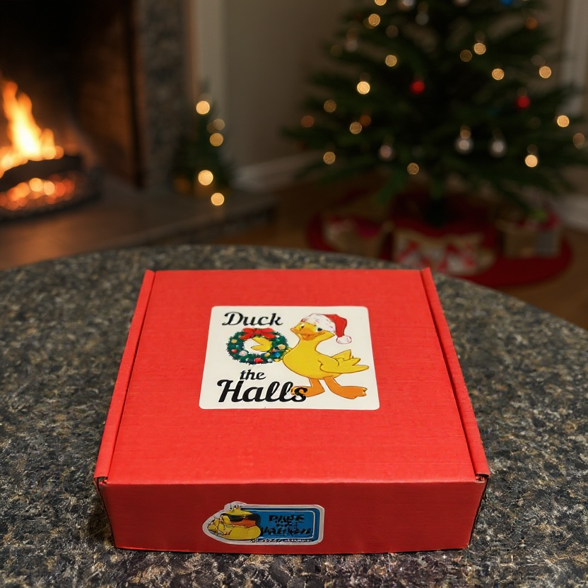 Red box with a Christmas-themed label on a table in front of a fireplace and decorated tree.