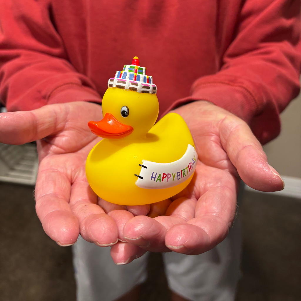 Yellow rubber duck with a birthday hat and 'Happy Birthday' banner held in hands.