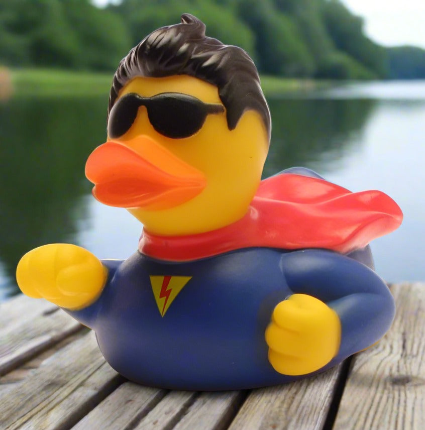 A toy rubber duck dressed in a superhero costume with a cape, wearing sunglasses, and positioned on a wooden surface with a body of water in the background.